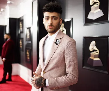 Zayn Malik standing on a red carpet in a pink suit, with framed Grammys symbols in the background.