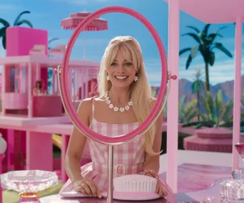 Barbie in checkered dress smiles into pink mirror, surrounded by pink decor and tropical background scene.