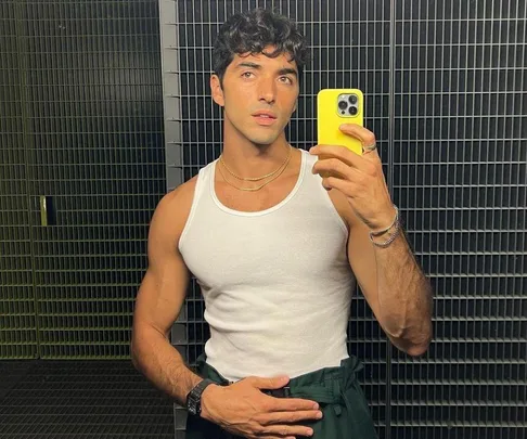 A man takes a mirror selfie in a white tank top, holding a yellow phone in a tiled bathroom.