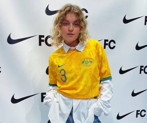 Person wearing a yellow Matildas jersey with number 3 over a white shirt, stands in front of a Nike and FC logo backdrop.