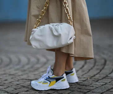 Sneakers with yellow accents paired with a beige dress and a white bag with a gold chain strap on cobblestone pavement.