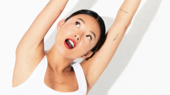 A woman with red lipstick and tattoos poses against a light background, looking upwards with arms raised.