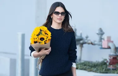 A woman in sunglasses holds a bouquet of sunflowers wrapped in brown paper, walking outdoors in a casual outfit.