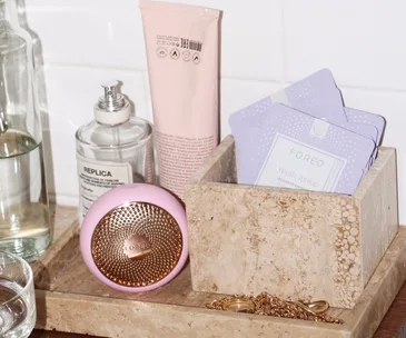 Skincare products on a bathroom counter: Foreo device, face masks, Replica bottle, pink tube, and gold jewelry.