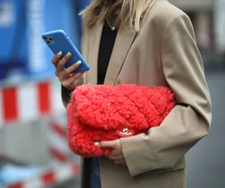 Woman in a beige coat holding a vibrant red fluffy clutch and a blue smartphone.