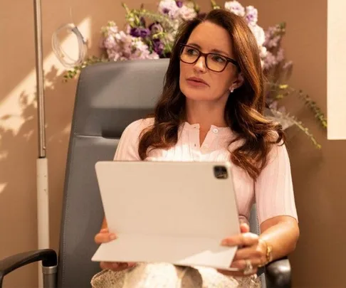 Woman with glasses holding a white tablet, seated in a chair, with flowers in the background.