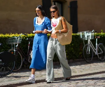 Two women walking outside, wearing sunglasses; one in a blue dress, the other in a tank top and wide pants.