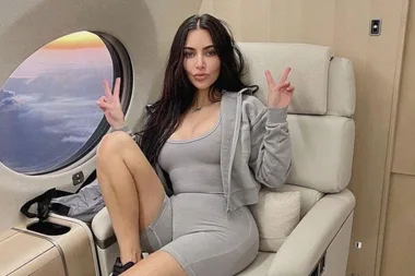 Person in a gray outfit sitting on a plane seat, posing with peace signs by a window showing a colorful sky.