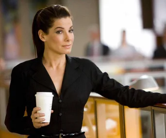 A woman in a black suit with a confident expression holds a coffee cup in an office setting.