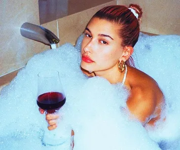 Person in a bubble bath holding a glass of red wine, looking relaxed.