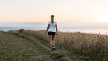 A person walking on a grassy path near the sea at sunset, showcasing solitude and nature.