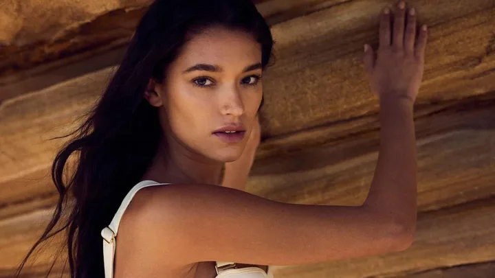 A woman with long dark hair in a white top stands against a rock formation, looking back over her shoulder.