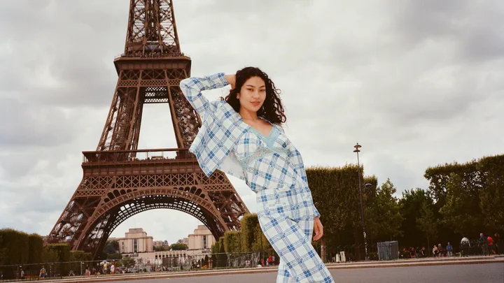 Woman posing in blue plaid outfit in front of the Eiffel Tower on a cloudy day.