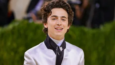 A young man with curly hair wearing a formal outfit smiles at an event with a green foliage background.