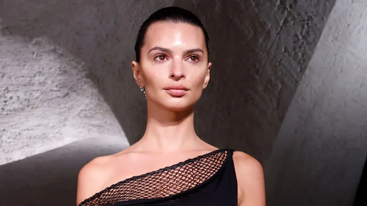 A person with minimal makeup, wearing a black dress with a mesh detail, and a slicked-back hairstyle against a textured wall.