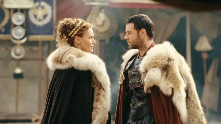 Scene from "Gladiator" with two characters in fur-trimmed cloaks facing each other in a Roman setting.