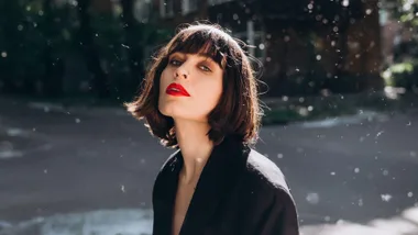 Woman with short brown hair and red lipstick in sunlight, wearing a black jacket, with blurry background.