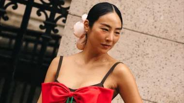 A woman with hair styled in a bun and wearing a red dress with floral decoration, standing against a stone wall.