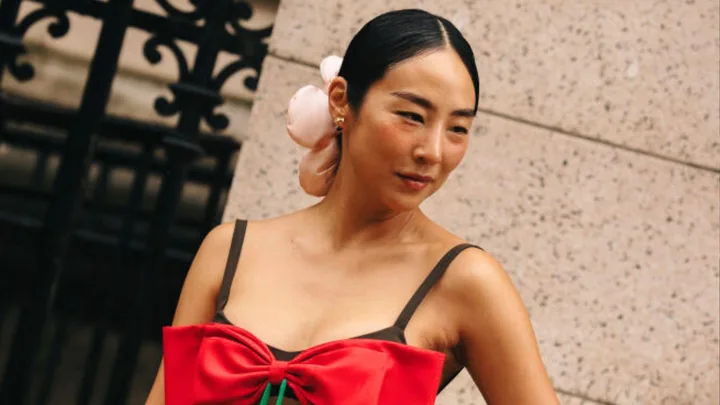 A woman with hair styled in a bun and wearing a red dress with floral decoration, standing against a stone wall.