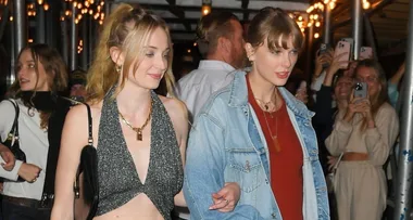 Two women walking arm in arm at night, one in a sparkly top, the other in a denim jacket, surrounded by people taking photos.