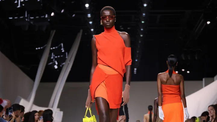 Model walks runway in orange asymmetric outfit, brown gloves, and red sunglasses, carrying a neon yellow bag at fashion show.