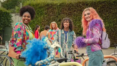 Three characters from "Sex Education" stand by bicycles, wearing colorful, eclectic outfits with bright smiles.