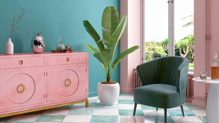 Stylish studio apartment with pink sideboard, green plant, teal chair, and checkerboard floor near a sunny window.