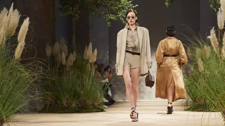 Model in beige outfit and sunglasses walks down a runway with lush greenery at Max Mara Spring/Summer 2024, Milan Fashion Week.