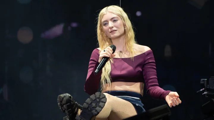 Singer with long blonde hair holds a microphone on stage, wearing a purple off-shoulder top and dark skirt.