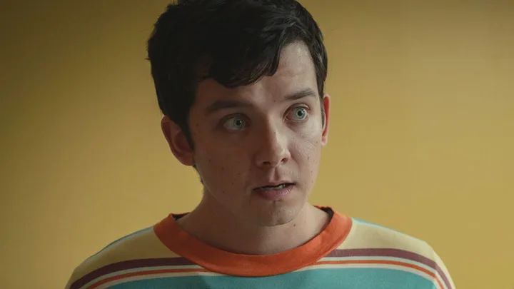 Young man with dark hair and a surprised expression, wearing a striped shirt, standing against a yellow background.