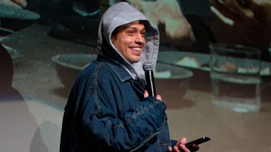 Comedian in a denim jacket and hoodie smiles while holding a microphone on stage.