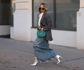 Woman in a denim maxi skirt, gray blazer, and silver boots, carrying a green handbag on a city street.