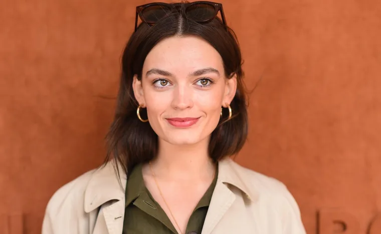 Woman with long brown hair, hoop earrings, trench coat, and sunglasses on head, smiling against a warm brown background.