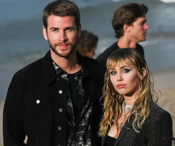 A couple stands on a beach, with the ocean in the background and people around them.