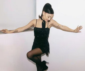 A woman in a sparkly black dress poses with arms outstretched, wearing black tights and heels, against a white background.