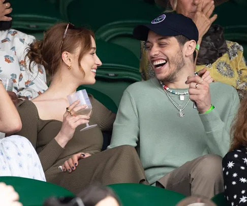 A couple smiling and laughing, seated in a stadium, one holding a drink, wearing casual outfits.