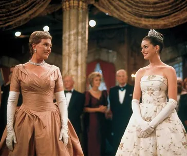 Two women in elegant gowns and gloves stand smiling at each other in a formal ballroom setting.