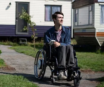 Character Isaac from "Sex Education" in a wheelchair, outdoors by trailers, smiling and wearing a blue jacket.