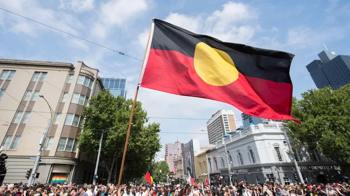 A large Aboriginal flag waves above a crowd during a street protest with city buildings in the background.