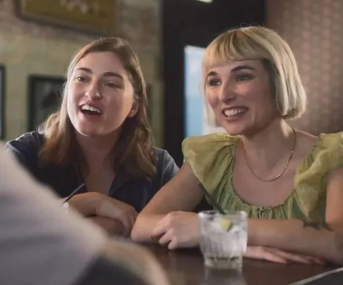 Two women smiling at a bar table, one with short blonde hair and a green top, the other with long brown hair.