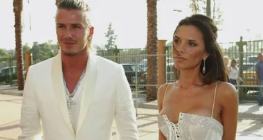 A couple in elegant white outfits at an outdoor event, with palm trees and a fence in the background.