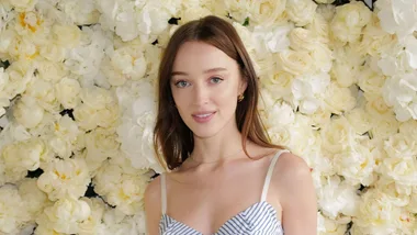 A woman with long brown hair poses in front of a wall of white roses, wearing a striped dress and gold earrings.