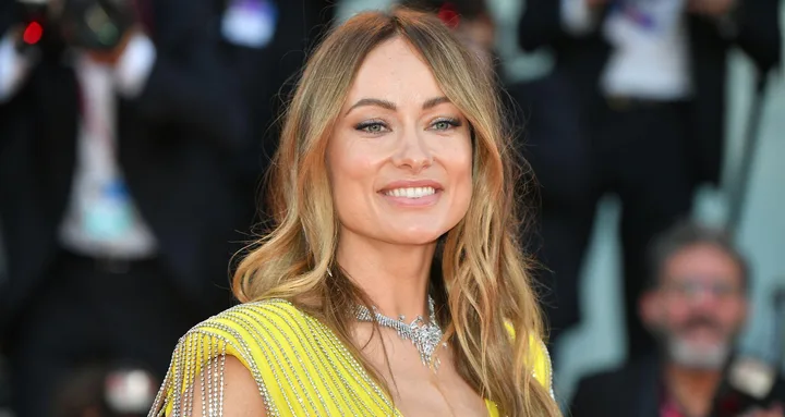 Woman in a yellow dress with wavy hair, smiling on a red carpet with photographers in the background.