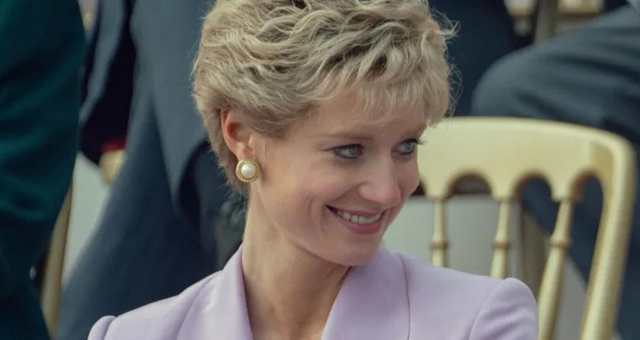 A smiling woman with short blonde hair wearing a lavender outfit and pearl earrings, seated outdoors.