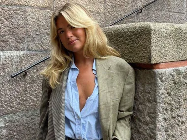 A woman with long blonde hair wearing a checkered blazer and blue shirt leans against a stone wall.