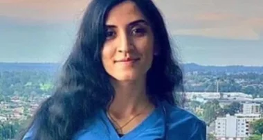 A woman with long black hair wearing a blue shirt stands outdoors with a cityscape in the background.