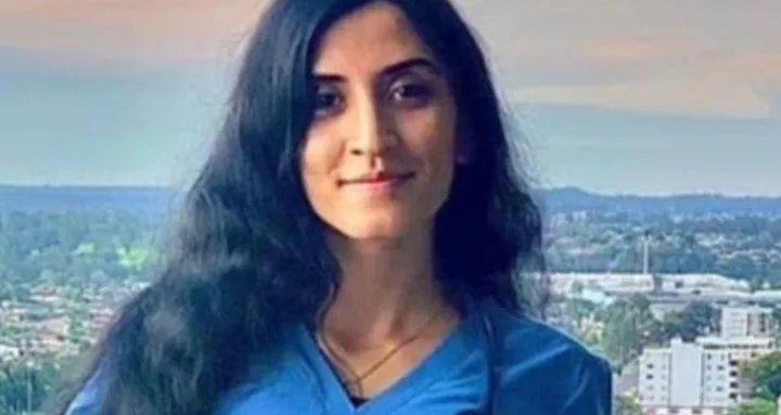A woman with long black hair wearing a blue shirt stands outdoors with a cityscape in the background.