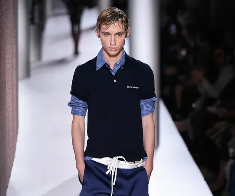 Model walks runway in navy polo over checked shirt and blue boardshorts with white ties, during Miu Miu fashion show.