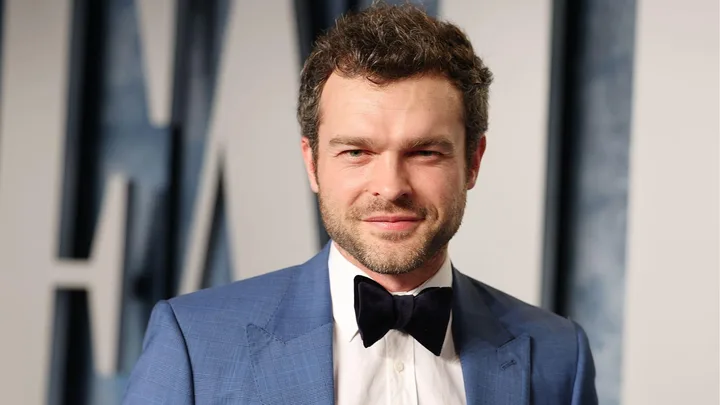 A man in a blue suit and bow tie is smiling at the camera against a blurred background.