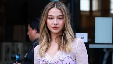 A woman with long blonde hair in a floral dress stands outside a building, looking directly at the camera.